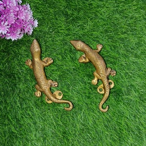 May include: Two gold-colored metal lizard door handles on a green artificial grass background.