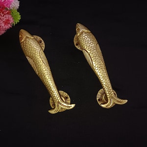 Pode incluir: Duas puxadores de porta dourados em forma de peixe, com designs detalhados de escamas e barbatanas. As puxadores estão posicionadas diagonalmente contra um fundo preto, sugerindo um tema decorativo ou artístico. Estes puxadores de peixe são provavelmente para um armário ou porta.