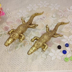 以下が含まれることがあります： 彫刻が施された金色のワニのドアノブが2つ。ノブは、ダイヤモンド模様の白い背景の上に置かれています。