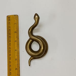 May include: A brass snake figurine, coiled in a stylized design. The snake is approximately 15 cm long, with a smooth, polished surface. The head is raised, and the body forms a loop. The figurine is set against a white background.