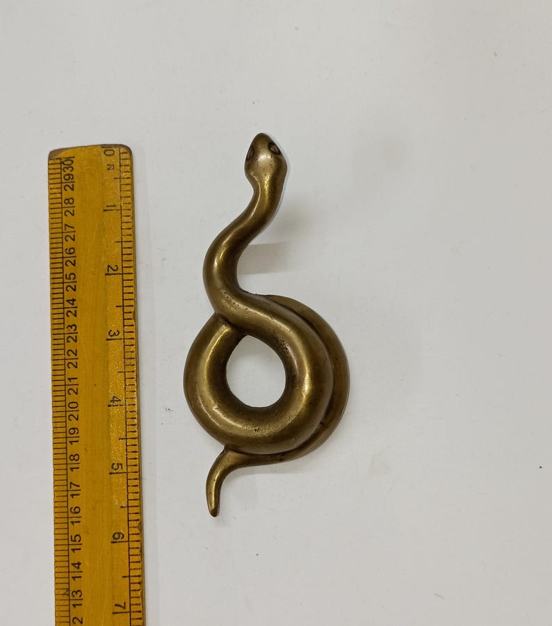 May include: A brass snake figurine, coiled in a stylized design. The snake is approximately 15 cm long, with a smooth, polished surface. The head is raised, and the body forms a loop. The figurine is set against a white background.