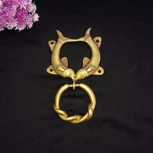 May include: A gold-toned brass door knocker in the shape of two kissing fish. The fish are detailed with scales and fins. The knocker has a circular ring for pulling.