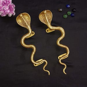 May include: A pair of gold-toned brass door handles shaped like cobras with intricate details. The handles are in a curled position, with the heads facing forward.