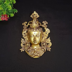 Puede incluir: Un colgante de pared de metal dorado que representa a una deidad con una expresión serena. La deidad está adornada con intrincados diseños florales y geométricos, creando una pieza visualmente atractiva y espiritual.