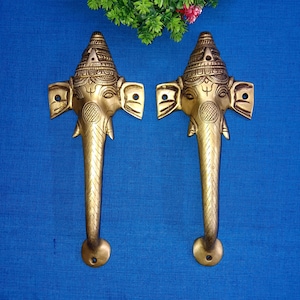 Puede incluir: Un par de tiradores de puerta de color dorado con un diseño de cabeza de elefante. Los tiradores están detallados con intrincados tallados y tienen un estilo indio tradicional.