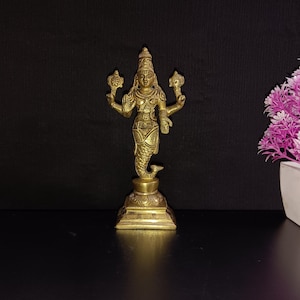 Puede incluir: Una estatua de metal dorado de una deidad hindú con cuatro brazos, sosteniendo objetos en cada mano. La estatua está de pie sobre una base cuadrada.