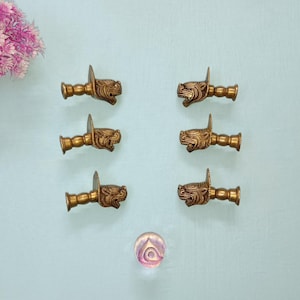 Puede incluir: Seis tiradores de cajón antiguos de color latón con un diseño detallado de cabeza de dragón. Los tiradores están dispuestos sobre una superficie azul claro, con un acento decorativo de vidrio y flores rosas.