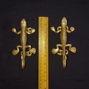 May include: Two gold-toned metal lizard hooks. The hooks are about 4 inches long and have a detailed lizard design. The hooks are mounted on a white background.