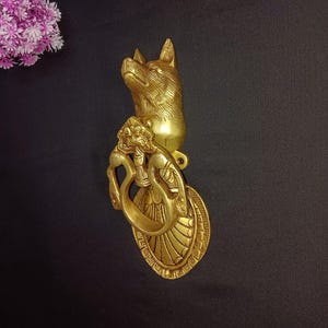 May include: A gold-toned door knocker featuring an intricate design of a fox's head and a decorative ring. The knocker is mounted against a black backdrop.