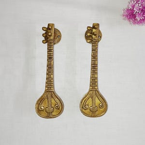 May include: Two gold-toned metal door handles shaped like sitar musical instruments. The handles feature intricate details and a traditional design.