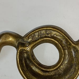 May include: A close-up of a bronze-coloured, abstract sculpture with a circular opening. The sculpture has a swirling, organic shape with a signature etched into the surface.