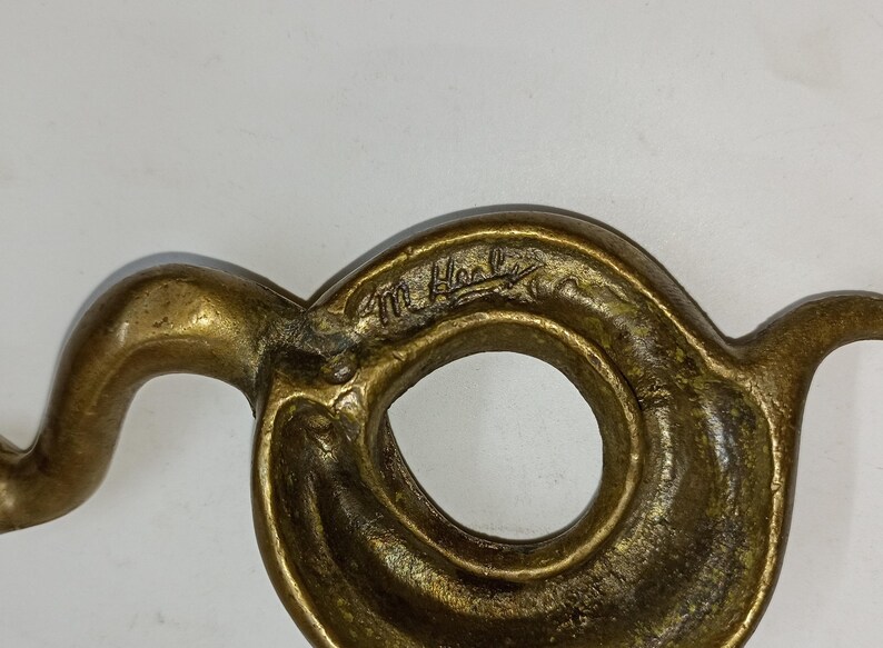 May include: A close-up of a bronze-coloured, abstract sculpture with a circular opening. The sculpture has a swirling, organic shape with a signature etched into the surface.