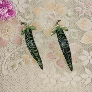 May include: Two decorative leaf-shaped metal handles. The handles are dark green with detailed vein patterns and gold-coloured accents on the leaf tips. The handles are designed to be mounted on a cabinet or drawer.