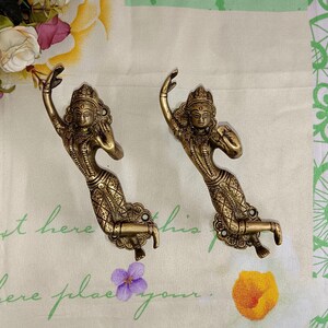 Op de afbeelding: Twee goudkleurige metalen haken in de vorm van vrouwen in traditionele Indiase kleding. De haken zijn gemonteerd op een witte stoffen achtergrond met een groen-wit bloemenpatroon.