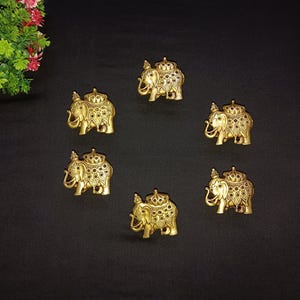 Könnte beinhalten: Sechs goldfarbene Elefantenfiguren mit aufwendigen Details sind auf einer schwarzen Oberfläche angeordnet. Jeder Elefant hat einen dekorativen Sattel und eine Krone. Es handelt sich wahrscheinlich um Dekorationsartikel oder Charms.