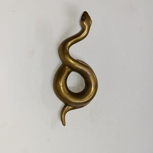 May include: A brass-toned snake sculpture in a coiled pose. The snake's head is raised, and its body forms a circular loop. The sculpture has a smooth, polished finish and is set against a white backdrop.