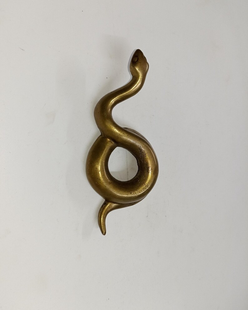 May include: A brass-toned snake sculpture in a coiled pose. The snake's head is raised, and its body forms a circular loop. The sculpture has a smooth, polished finish and is set against a white backdrop.