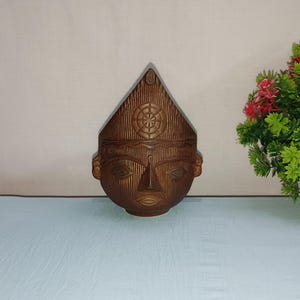 May include: A wooden carved sculpture of a face with a pointed hat. The sculpture is brown and has intricate details.