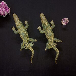 Peut inclure: Deux poignées de porte en laiton en forme d'alligator de style antique. Les poignées de porte sont vertes et dorées avec une surface texturée. Les poignées de porte en alligator sont posées sur une surface noire. Un petit bouquet de fleurs violettes est dans le coin supérieur gauche.