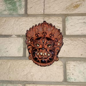 May include: A red and black ceramic mask with intricate details and a fierce expression. The mask is hanging on a white brick wall.