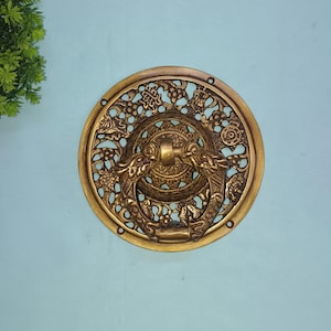 Op de afbeelding: Osiers messing deurklopper met een cirkelvormig ontwerp. De klopper heeft een centrale ring met twee drakenkoppen en ingewikkelde bloemen- en geometrische patronen. Het messing heeft een warme, verouderde afwerking.