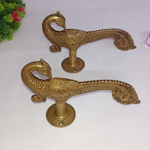 May include: Two brass peacock-shaped door handles. The handles are gold-colored with detailed feather and body patterns. Each handle has a circular base and a curved, elongated body. The peacock's head is at the top, and the tail feathers fan out.