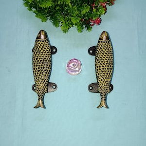 May include: Two brass fish door handles with a yellow and black finish. The handles are shaped like fish with scales and fins.