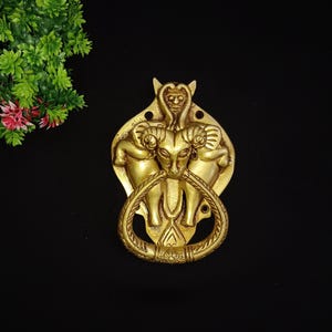 May include: A gold-toned door knocker with a detailed design of two rams flanking a central figure. The knocker has a ring-shaped handle.