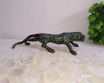 May include: A black and green spotted leopard figurine. The sculpture is in a stalking pose, with its body low to the ground. The leopard's spots are a bright green color, and the figurine is made of a dark, possibly metallic material.