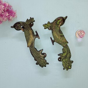 Może przedstawiać: Dwa ozdobne, mosiężne uchwyty do drzwi w kształcie pawia, z zielonymi akcentami. Uchwyty są szczegółowo zdobione skomplikowanymi wzorami i umieszczone na jasnoniebieskim tle. Obecna jest również mała, przezroczysta, dekoracyjna szklana kulka.