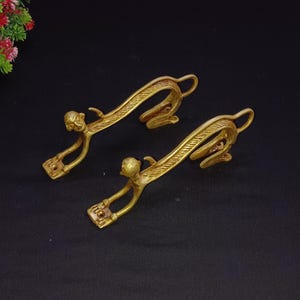 May include: Two golden-colored, stylized monkey-shaped drawer pulls. Each pull features a monkey figure with outstretched arms and a curved tail. The pulls are made of metal and are set against a dark background.