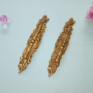 以下が含まれることがあります： 複雑な花の模様が施された、金色の金属製の引き出しの取っ手が2つ。取っ手には装飾的な隆起した中央部分があります。