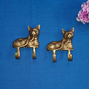 Può includere: Due ganci in metallo dorato a forma di gatto con un dettaglio a fiocco. I ganci sono montati su una superficie blu.