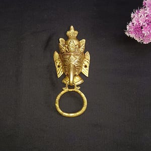May include: A gold door knocker with an ornate design featuring a face with an elephant trunk. The knocker is attached to a circular ring.
