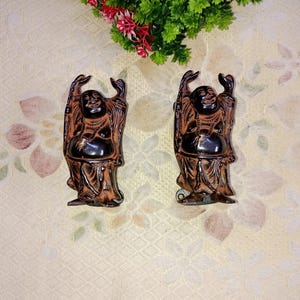 May include: Two brown and black laughing Buddha figurines with raised arms. The figurines are standing on a white surface.