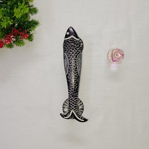 May include: A black and white fish-shaped door handle with detailed scale and fin patterns. The handle is made of a dark material and has a decorative design. A small, iridescent, round object is also present.