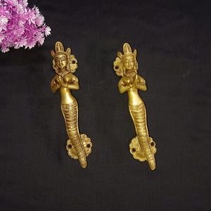 以下が含まれることがあります： 祈りのポーズをした人魚のような姿の真鍮製ドアハンドル2つ。 金色のハンドルには、王冠や鱗のある尾など、細部までこだわった装飾が施されています。 背景は黒で、左上にはピンクの花が咲いています。