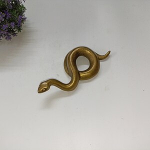 May include: A bronze snake ornament in a coiled pose. The snake has a detailed head and body, with a smooth, polished finish. The ornament is set against a white background, with a hint of purple flowers in the upper left corner.