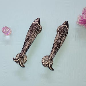 May include: Two black and white fish-shaped door handles with a detailed scale pattern. They are set against a light blue background. A pink flower and a clear, iridescent gem are also in the image.