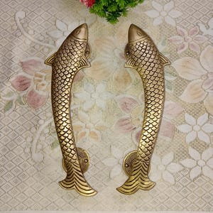 Könnte beinhalten: Ein Paar goldfarbene, fischförmige Türklinken. Die Griffe sind mit Schuppen verziert und haben eine gebogene Form.