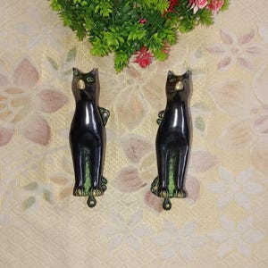 Könnte beinhalten: Zwei schwarze Katzen-Türgriffe aus Metall mit grünen Augen und goldenen Akzenten. Die Griffe befinden sich auf einem floral gemusterten Hintergrund.