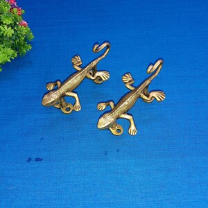 May include: Two brass lizard-shaped door handles. The handles are detailed with textured scales and curled tails. The lizards are positioned on a blue surface, with a green plant in the upper left corner.