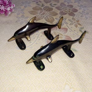 May include: Two black metal dolphin door handles with gold accents. The dolphins are facing the right side of the image. The handles are mounted on a white surface.