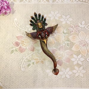 May include: A decorative door handle made of brass with a green, red and gold finish. The handle is shaped like a snake with a human figure on its back. The human figure has a halo and is holding a flower in its hand.