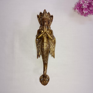 Peut inclure: Une poignée de porte en métal doré en forme de sirène avec des ailes. La sirène est en position de prière, les mains jointes. La poignée est ornée et détaillée.