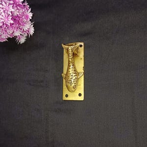 May include: A gold-colored brass door knocker shaped like a fish with detailed scales. The knocker is mounted on a rectangular plate with two holes for screws.