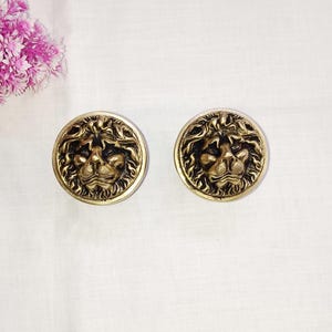 Könnte beinhalten: Zwei goldfarbene Löwenkopfknöpfe mit schwarzen Details. Die Knöpfe sind rund und haben ein erhabenes Design.