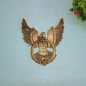 Puede incluir: Un llamador de puerta de metal dorado con forma de pájaro con las alas extendidas. El pájaro sostiene un anillo en su pico.