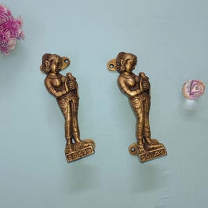 Könnte beinhalten: Zwei Türgriffe aus Messing in Form einer stehenden Figur, die einen Gegenstand hält. Die Griffe weisen aufwendige Details auf und sind auf einer hellblauen Oberfläche montiert. Ein rosa Blumenarrangement und eine dekorative Glaskugel sind ebenfalls vorhanden.