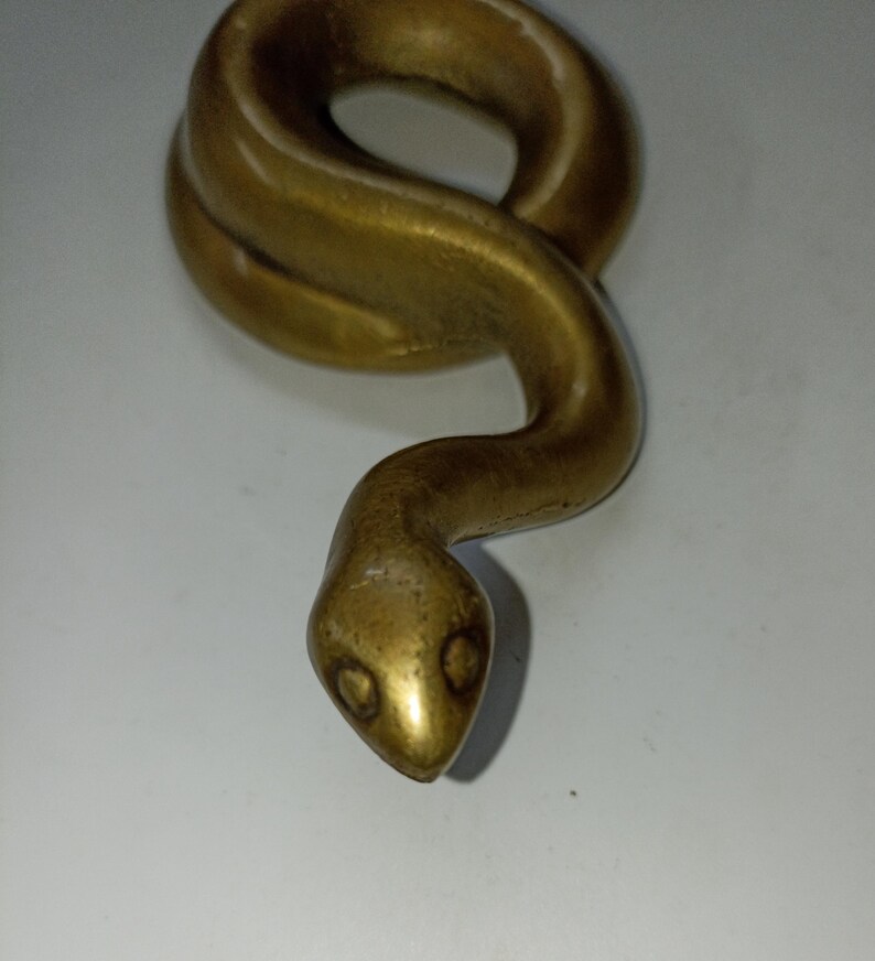 May include: A brass-coloured snake sculpture. The serpent is coiled with a detailed head and visible eye sockets. The sculpture has a smooth, polished finish against a white background.